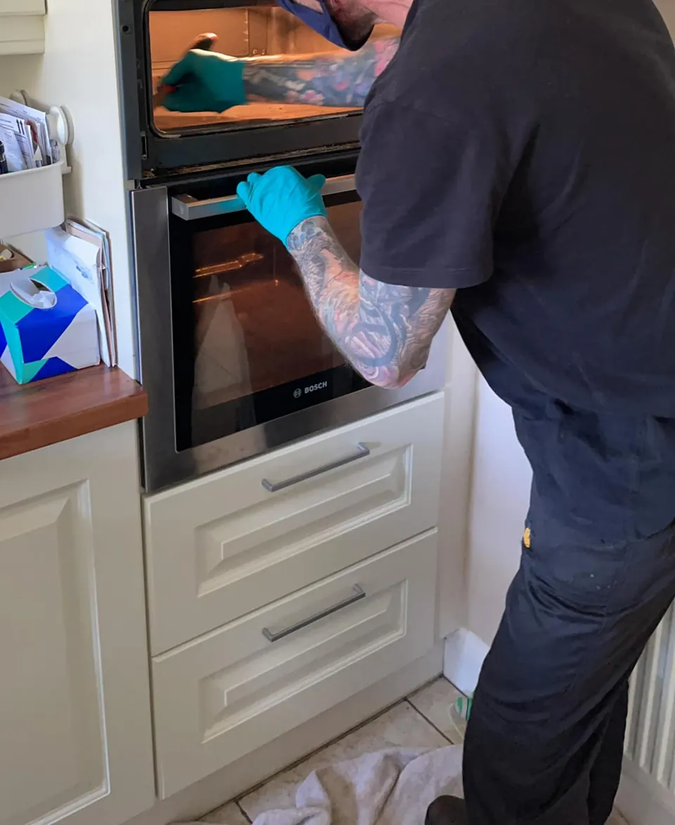 A trainee in gloves and a face mask cleaning inside a built-in oven in a customer's kitchen